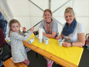 Das Foto zeigt Ulrike Gräf und ihre Tochter Madeleine, die für Kinder und Jugendliche das Bemalen von Tassen anbieten. Ein junges Mädchen bearbeitet gerade eine Tasse und blickt interessiert in die Kamera.