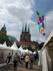 Auf dem Foto blickt man in eine der Zeltgassen auf dem Domplatz. Menschen bewegen sich dicht durch die Gasse. Im Hintergrund ragt der Dom über dem Domplatz empor.